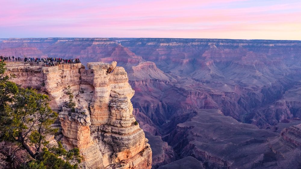 A pink sunset pours over a crowd of visitors enjoying the canyon views from Mather Point in Grand Canyon National Park, Arizona