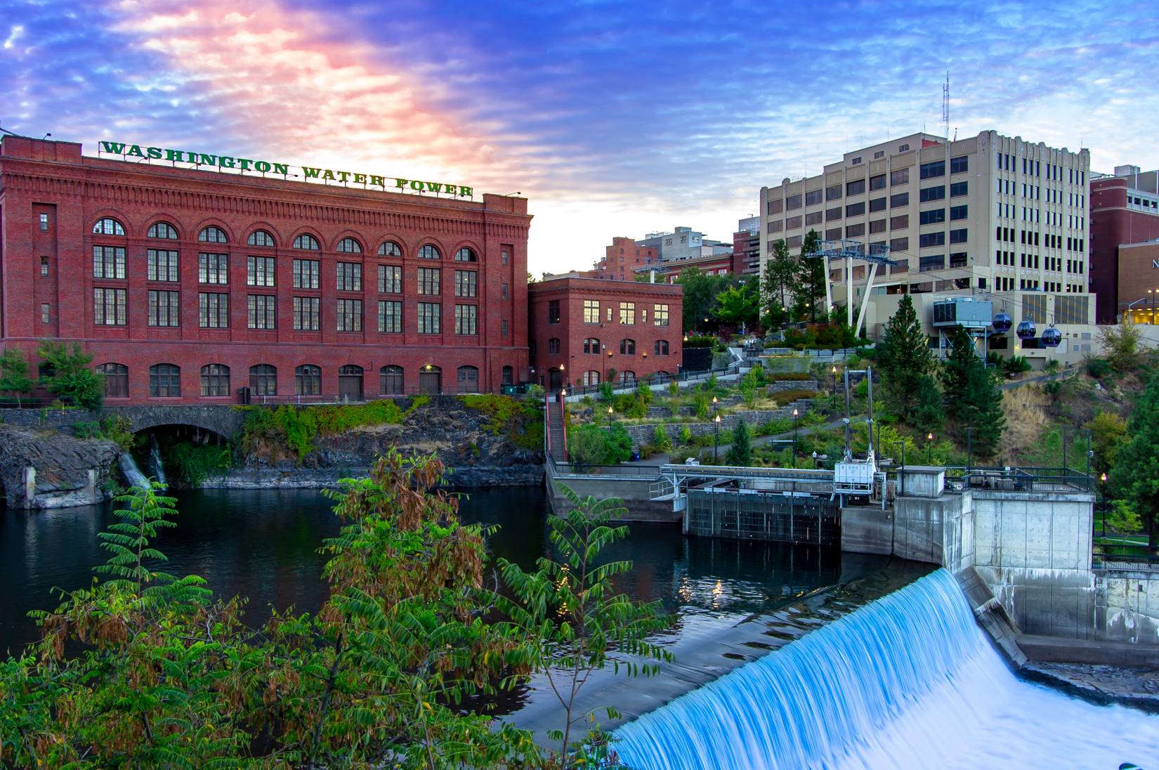 A view of the Spokane River Dam against the backdrop of the downtown skyline at sunrise. 