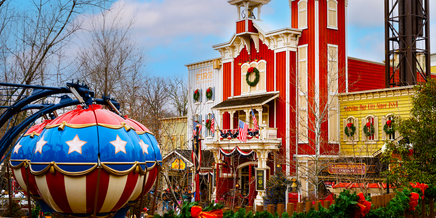Red, white a blue theme park ride, a red old-fashioned building with Christmas wreath decorations, and bare winter trees in the distance on a cloudy day