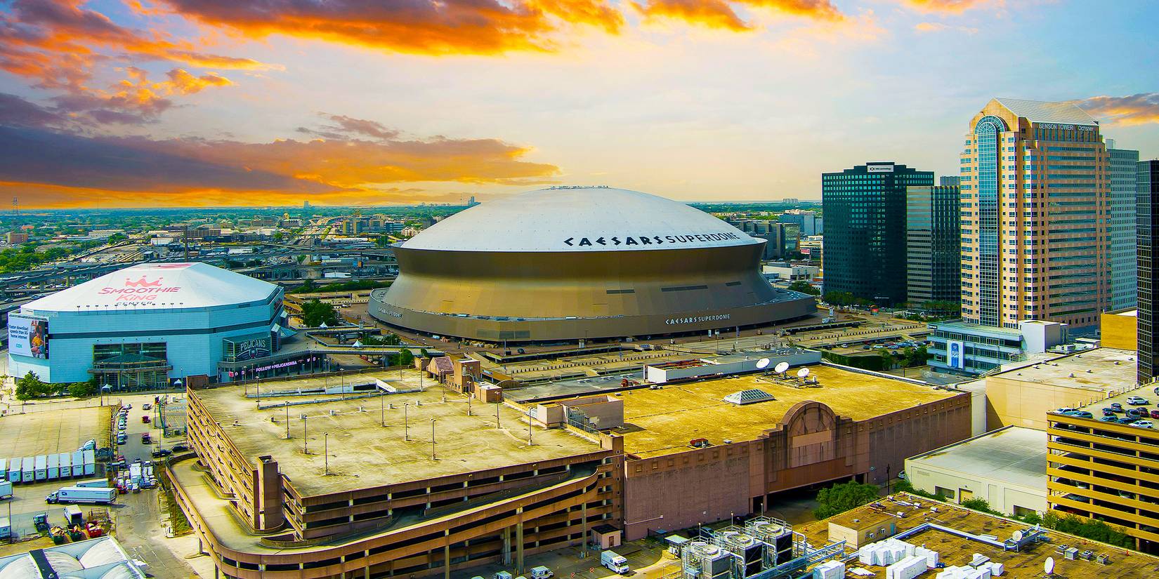 Sunset over a city with skyscrapers, a large white-roofed sports stadium and warehouses in the foreground