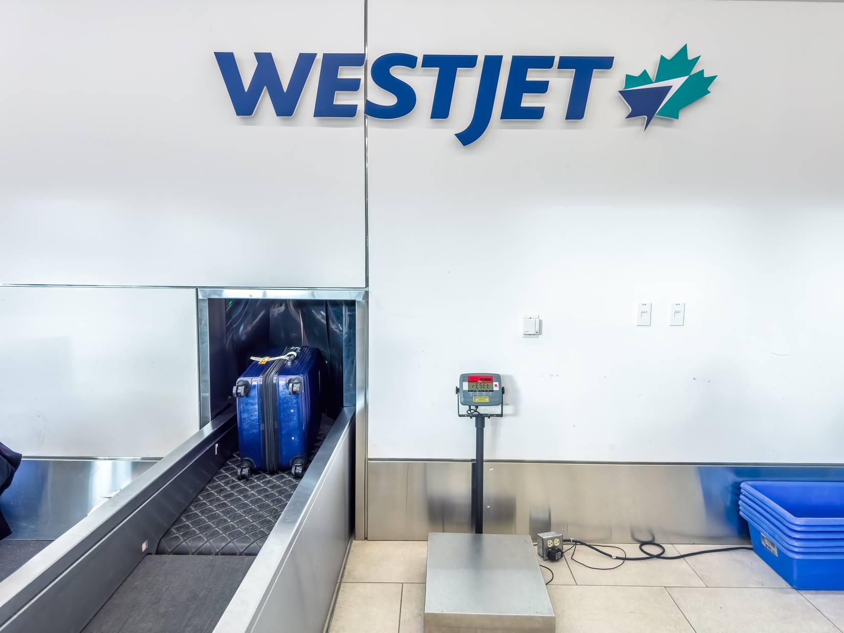 A blue suitcase is being loaded onto a conveyor belt at a WestJet check-in counter. The airline's logo is prominently displayed above.