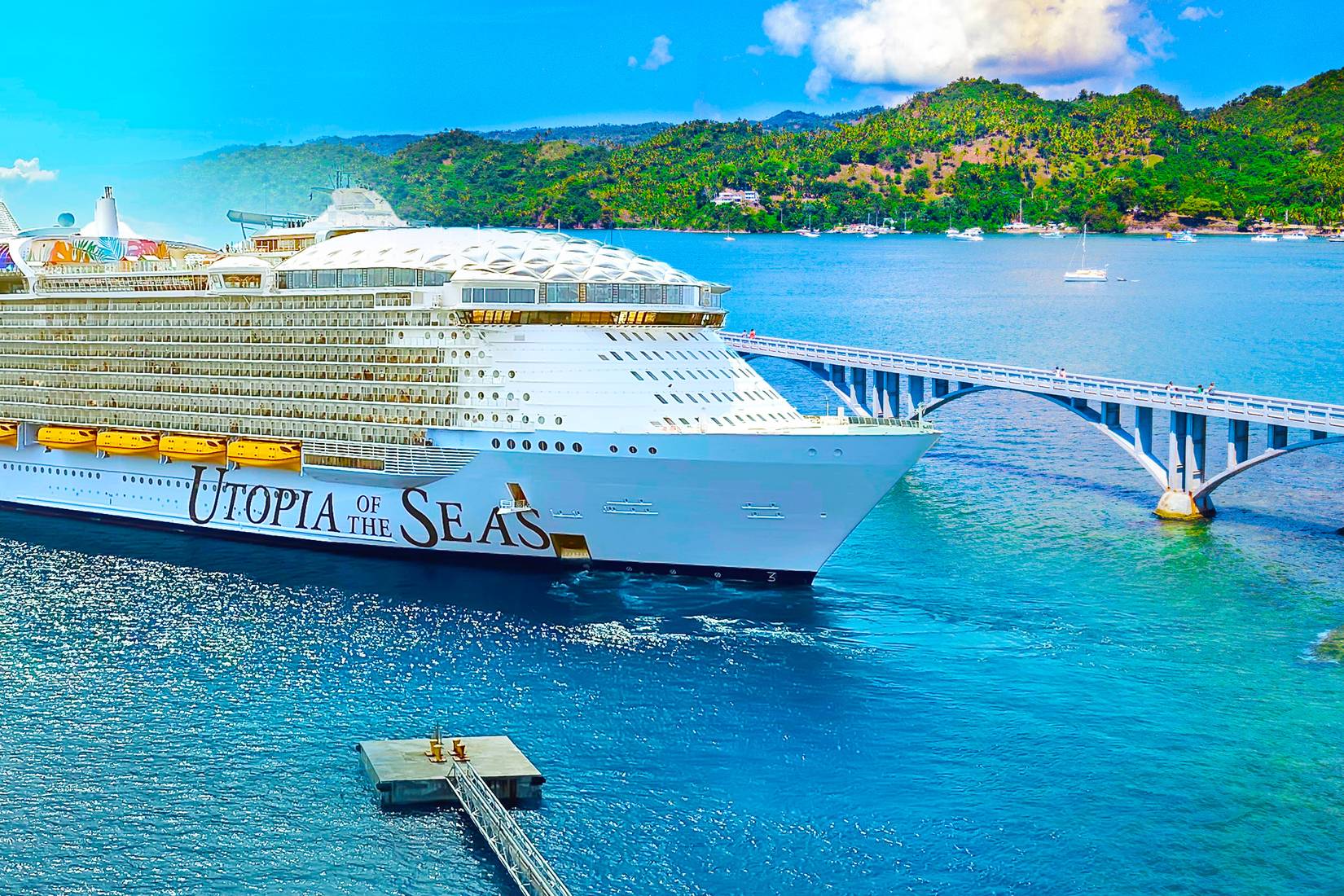White Caribbean Cruse ship on clear blue water on the left side of the image, the coast of Samana with a long bridge, sailboats and green hills on the right side of the image