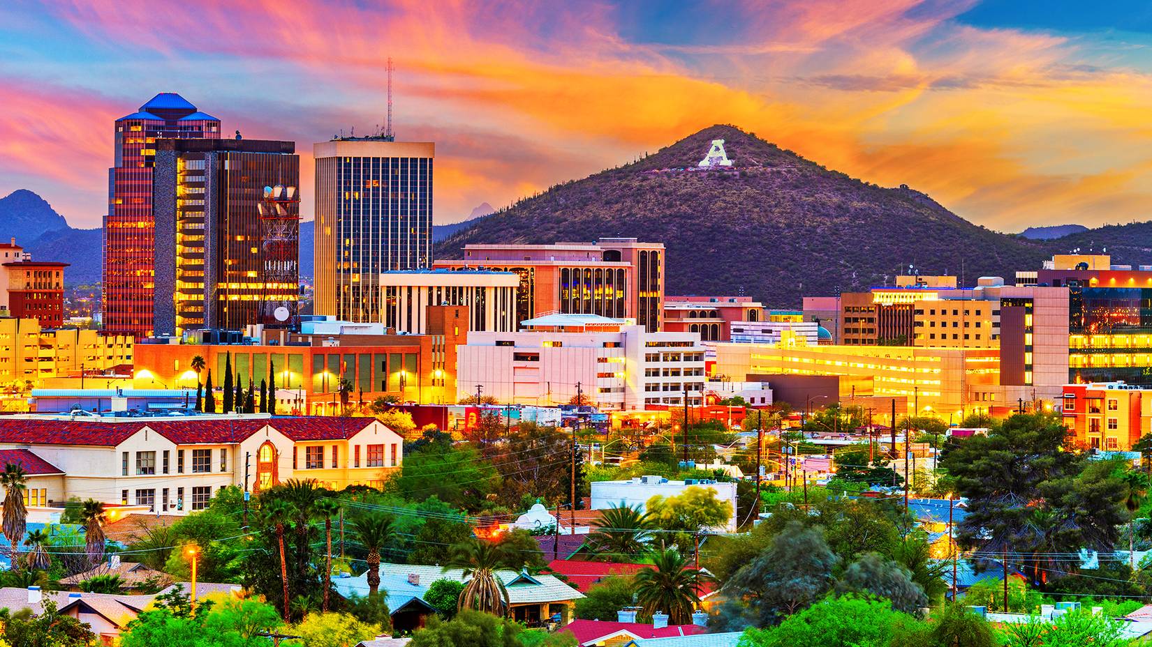Green trees, between a mix of old fashioned buildings and skyscrapers lit with orange light at sunset with mountains in the distance