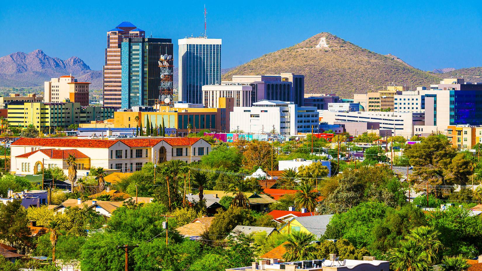 Photo of Tuscon Arizona with green trees and palms, some old-fashioned red roofed buildings and skyscrapers with a mountain in the distance